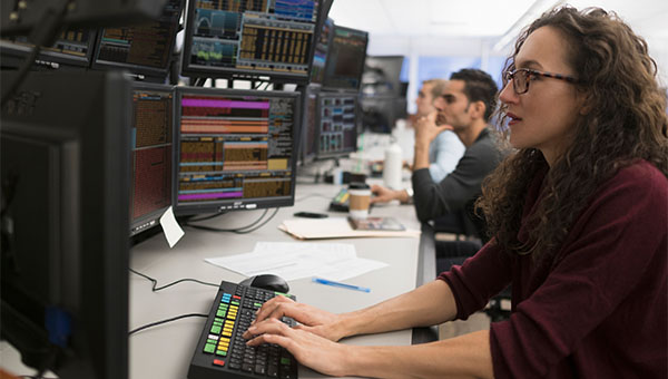 Employee monitors multiple data dashboards on computer screens in an office while colleagues work in the background