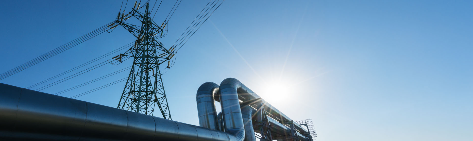 Large industrial pipelines beneath electrical transmission lines against a bright blue sky