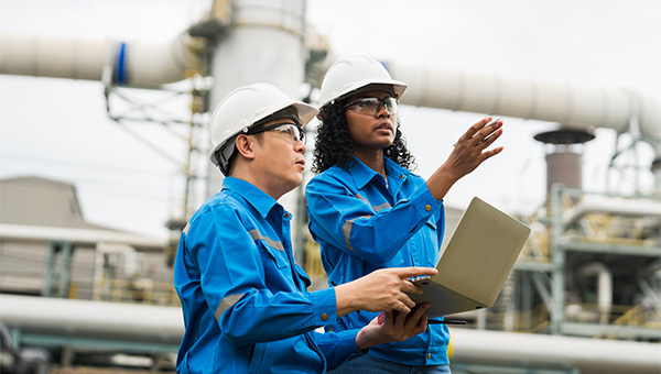 Two workers in blue uniforms and hard hats review a laptop while discussing equipment at an industrial facility