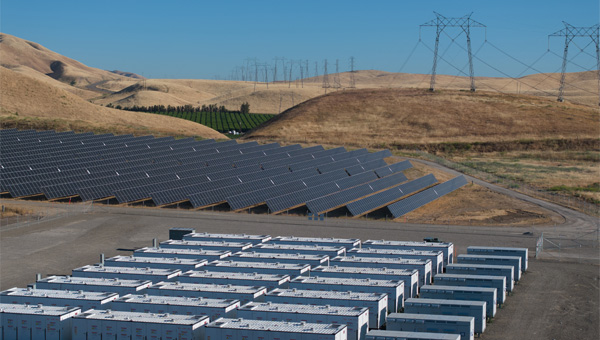 Battery storage units beside a large solar panel array with power transmission lines across rolling hills