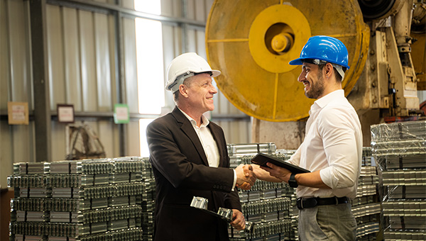 Two workers wearing hard hats shake hands in a manufacturing facility while reviewing a tablet