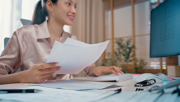 Person holding documents while working at a desk with a computer, keyboard, and office paperwork