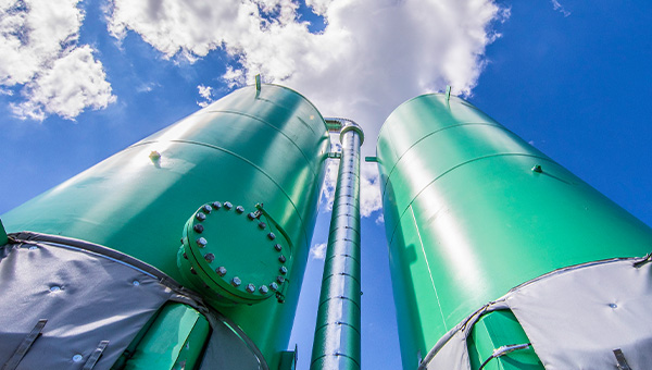 Low-angle view of two large green industrial tanks and a metal pipe against a bright blue, cloudy sky.